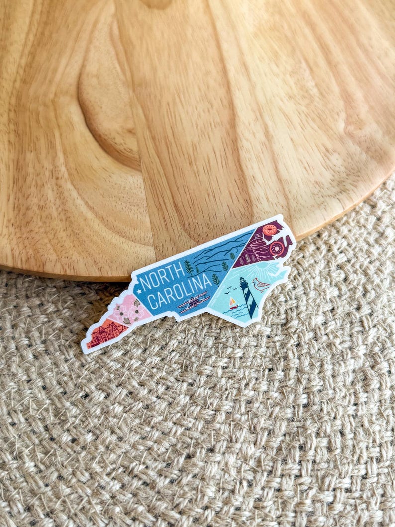 May include: A white sticker with a colorful outline of the state of North Carolina. The sticker features various illustrations of landmarks and natural features, including a lighthouse, mountains, and a beach scene. The text "NORTH CAROLINA" is printed in black letters on the sticker.