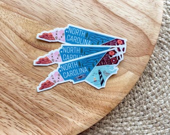 May include: Four North Carolina state outline stickers with a blue background and colourful illustrations of landmarks and nature. The stickers are stacked on top of each other on a wooden surface.