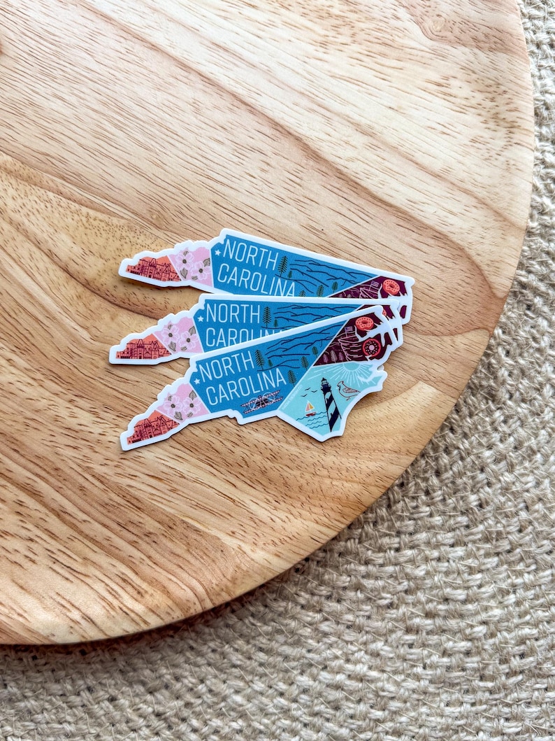 May include: Four North Carolina state outline stickers with a blue background and colorful illustrations of landmarks and nature. The stickers are stacked on top of each other on a wooden surface.
