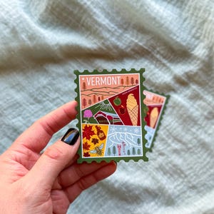 May include: A green sticker with a Vermont theme, featuring illustrations of a landscape, an apple, an ice cream cone, and other Vermont symbols. The word "VERMONT" is at the top. The sticker is held by a hand.