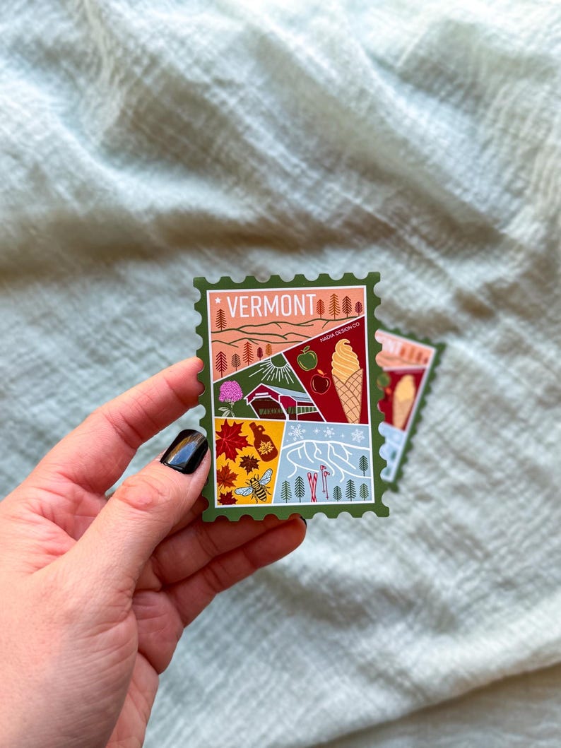 May include: A green sticker with a Vermont theme, featuring illustrations of a landscape, an apple, an ice cream cone, and other Vermont symbols. The word "VERMONT" is at the top. The sticker is held by a hand.