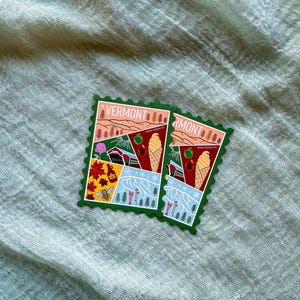 May include: Two Vermont-themed stickers with a postage stamp design. Each sticker features illustrations of a landscape with trees, a farm, ice cream cones, and the word "VERMONT". The stickers have a green border and are set against a light green fabric.