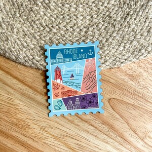 Può includere: Un adesivo azzurro con un design a francobollo con illustrazioni di monumenti del Rhode Island. L'adesivo include un faro, un ponte e il Campidoglio. Il testo "RHODE ISLAND" è in alto.