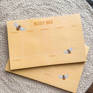 May include: Two yellow weekly planner pads with a bee illustration and the text "BUSY BEE" at the top. The days of the week are listed across the top of the pad, with Monday, Tuesday, Wednesday, Thursday, Friday, Saturday, and Sunday listed.