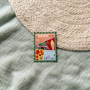 May include: A green Vermont-themed sticker with a postage stamp design. The sticker features illustrations of landscapes, an apple, an ice cream cone, a covered bridge, and the word "VERMONT" at the top.