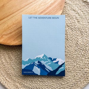 May include: A blue and white notepad with a mountain range illustration and the text "LET THE ADVENTURE BEGIN". The notepad has a dotted pattern and the text "NACIA DESIGN CO" at the bottom.