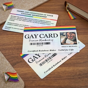 Two personalized 'GAY CARDs' stacked, one showing a custom photo and name 'SHAUN MICHAEL', on a light textured surface with a small pride flag pin and a book with a rainbow bookmark in the background.