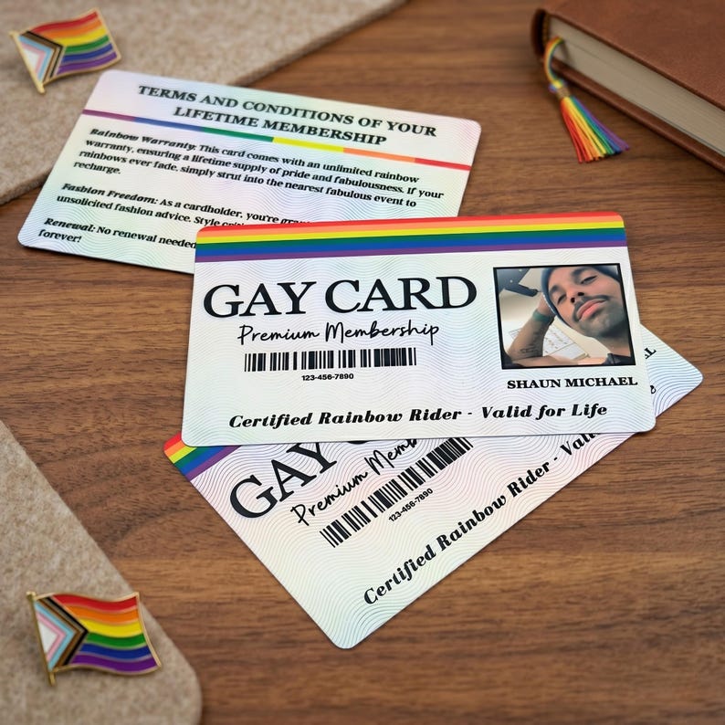 Two personalized 'GAY CARDs' stacked, one showing a custom photo and name 'SHAUN MICHAEL', on a light textured surface with a small pride flag pin and a book with a rainbow bookmark in the background.