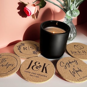 Puede incluir: Cinco posavasos de corcho con texto personalizado y fechas. Los posavasos presentan una variedad de diseños, incluyendo iniciales, anillos de boda y nombres. Los posavasos están dispuestos alrededor de una vela negra.