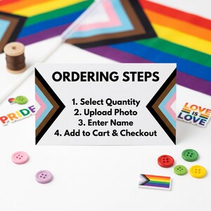 A white card titled 'ORDERING STEPS' with instructions to 'Select Quantity, Upload Photo, Enter Name, Add to Cart & Checkout' surrounded by small rainbow Pride flags and colorful buttons on a white surface.