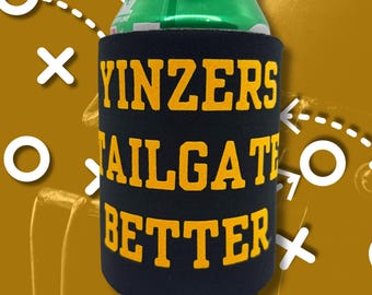 May include: A black drinks can cooler with yellow text reading "YINZERS TAILGATE BETTER." A green can is partially visible at the top. The background is a golden-brown colour with white football play diagrams.