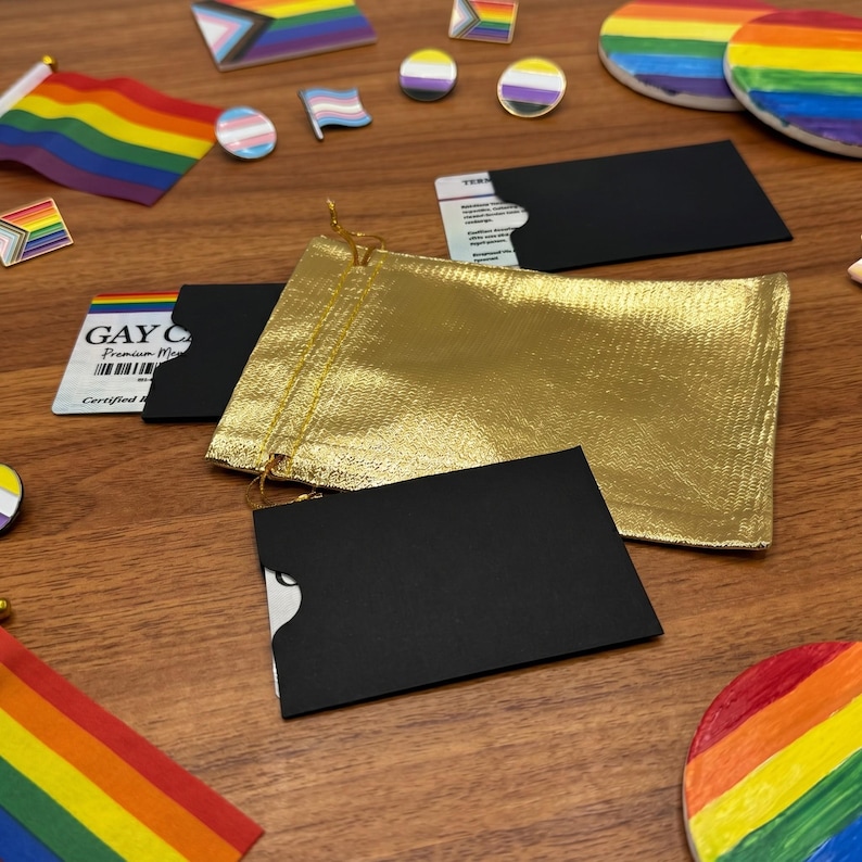 Overhead shot showing a personalized 'GAY CARD' next to small black and gold envelopes, various LGBTQ+ pride pins, and small rainbow flags on a wooden table.