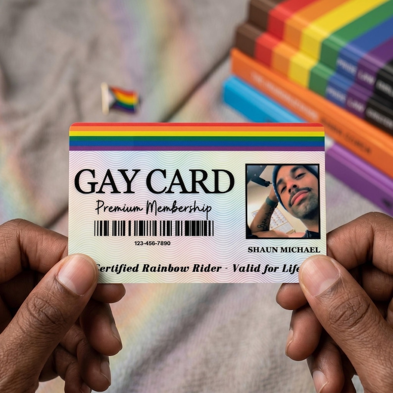 Two hands holding a personalized 'GAY CARD' with a photo of a man and the name 'SHAUN MICHAEL'. Rainbow colored books are blurred in the background, signifying an LGBTQ+ theme.