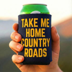 May include: A navy blue can cooler with the text "TAKE ME HOME COUNTRY ROADS" in yellow. The cooler is wrapped around a green can. The image is of a person holding the can.