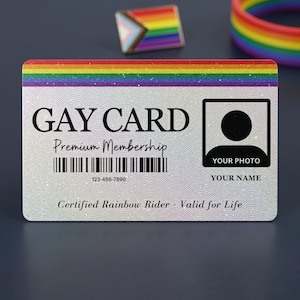 Front view of a custom 'GAY CARD Premium Membership' featuring a rainbow stripe, a glittery background, barcode, 'YOUR PHOTO' placeholder, and 'YOUR NAME' placeholder. 'Certified Rainbow Rider - Valid for Life' is printed below. A pride pin and rainbow bracelet are in the background.