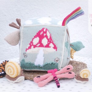 May include: A handmade crochet cube toy with a mushroom design, a rainbow, and other attached crochet elements. The cube is light blue with a pink and white mushroom, green grass, and a rainbow. Additional crochet toys include a dragonfly and snails.