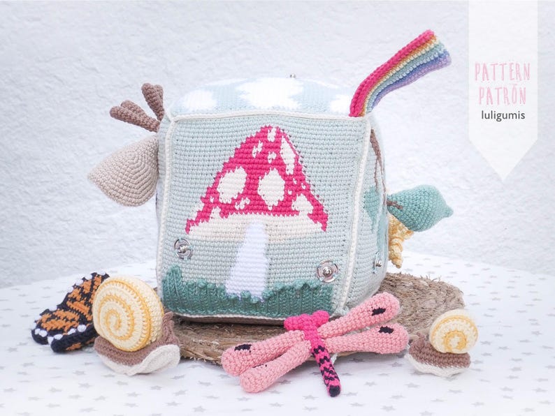 May include: A handmade crochet cube toy with a mushroom design, a rainbow, and other attached crochet elements. The cube is light blue with a pink and white mushroom, green grass, and a rainbow. Additional crochet toys include a dragonfly and snails.