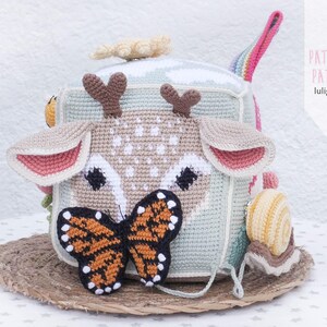 May include: A handmade crochet cube toy featuring a deer face with brown antlers, pink-lined ears, and a butterfly. The cube has a light green and beige color scheme, with a rainbow handle and a snail toy attached. The text "PATTERN PATRON luligumis" is visible.