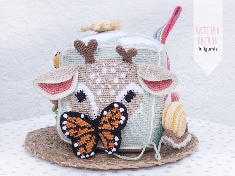 May include: A handmade crochet cube toy featuring a deer face with brown antlers, pink-lined ears, and a butterfly. The cube has a light green and beige color scheme, with a rainbow handle and a snail toy attached. The text "PATTERN PATRON luligumis" is visible.