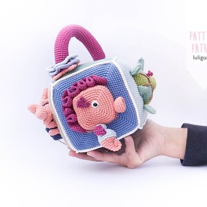 May include: A crocheted cube toy with a handle. Each side of the cube features a different animal, including a crab, a fish, a turtle, and a starfish. The toy is made in shades of blue, pink, green, and white.