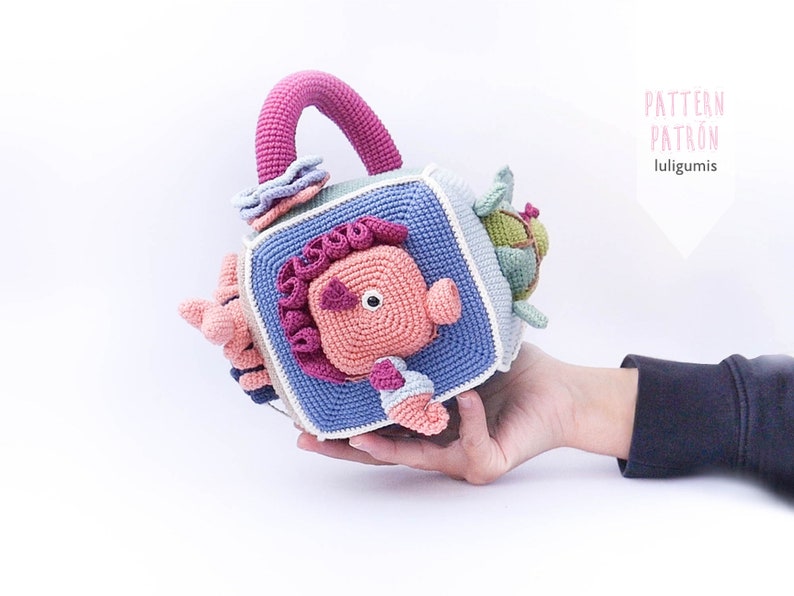 May include: A crocheted cube toy with a handle. Each side of the cube features a different animal, including a crab, a fish, a turtle, and a starfish. The toy is made in shades of blue, pink, green, and white.