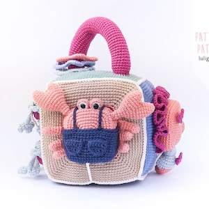 May include: A crocheted cube toy with a pink handle, featuring a crab, a fish, and other sea creatures. The cube is made of different colored squares and has a white border. The toy is designed for babies and toddlers.