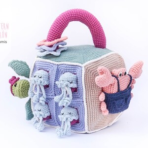 May include: A crocheted cube toy with a pink handle, featuring a turtle, a crab, and multiple jellyfish. The cube is made of different colored squares and has a variety of textures.