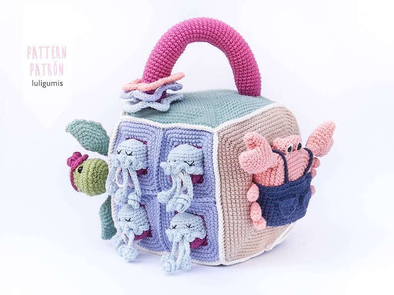 May include: A crocheted cube toy with a pink handle, featuring a turtle, a crab, and multiple jellyfish. The cube is made of different colored squares and has a variety of textures.