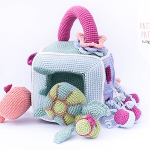May include: A crocheted toy set with a blue and white cube with a handle, a green turtle, a pink seahorse, and pink jellyfish. The cube has a hole in the front and the turtle fits inside. The set is labeled "PATTERN PATR&Oacute;N luligumis".
