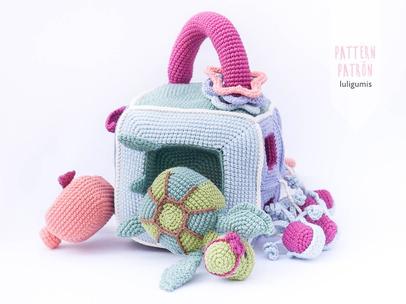 May include: A crocheted toy set with a blue and white cube with a handle, a green turtle, a pink seahorse, and pink jellyfish. The cube has a hole in the front and the turtle fits inside. The set is labeled "PATTERN PATR&Oacute;N luligumis".