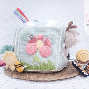 May include: A crocheted baby toy cube in light blue, featuring a large pink flower, green grass, and a rainbow handle. Additional crocheted toys include a bee, sun, snail, and butterfly. The cube sits on a woven mat. Text on the right says "PATTERN PATRON luligumis".