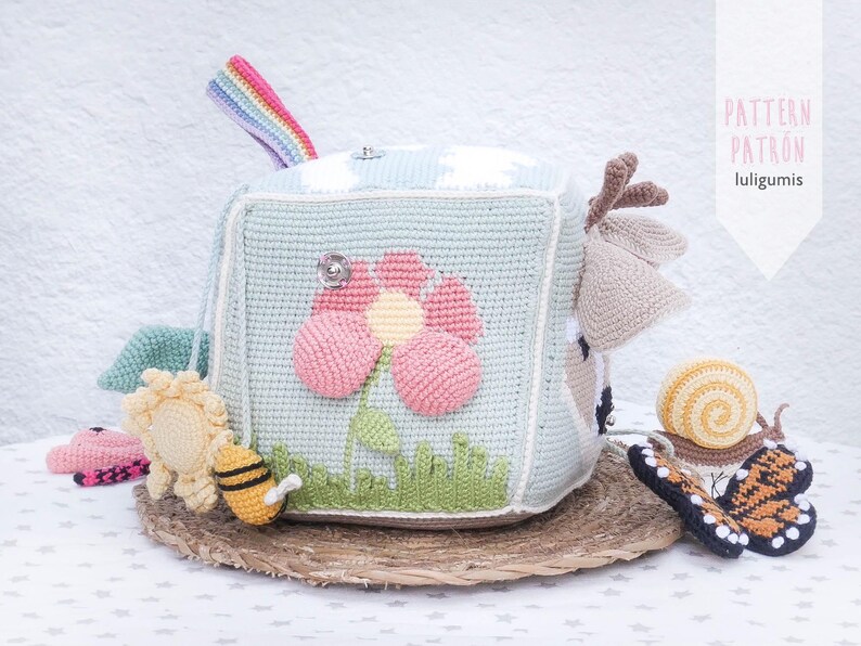 May include: A crocheted baby toy cube in light blue, featuring a large pink flower, green grass, and a rainbow handle. Additional crocheted toys include a bee, sun, snail, and butterfly. The cube sits on a woven mat. Text on the right says "PATTERN PATRON luligumis".