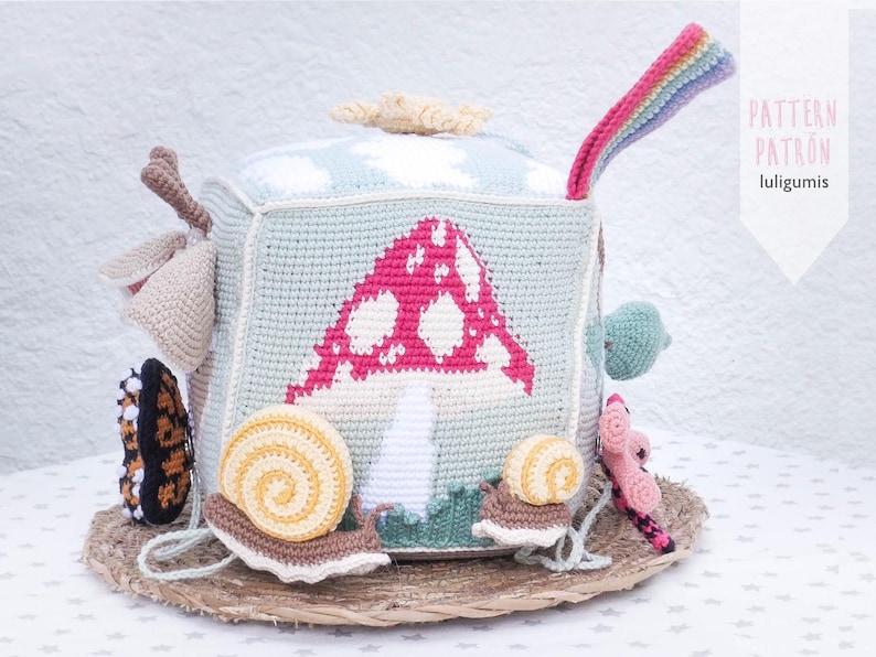 May include: A handmade crochet cube toy with a mushroom design in pink and white. The cube features various attached crochet elements, including a rainbow, snails, and a butterfly. The cube is placed on a woven mat. The text "PATTERN PATRON luligumis" is visible.