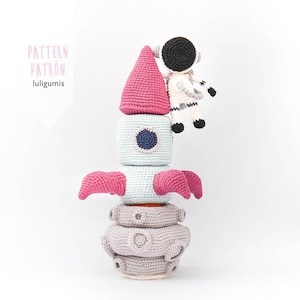 Puede incluir: Un juguete cohete de crochet con una parte superior rosa, un cuerpo azul y una base gris. Una pequeña figura de astronauta de crochet está sentada en la parte superior del cohete.