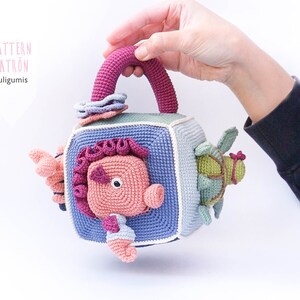 May include: A crocheted cube toy with a handle, featuring a pink seahorse, a green turtle, and a blue and white flower. The cube is made of different colored yarn and has a textured surface.