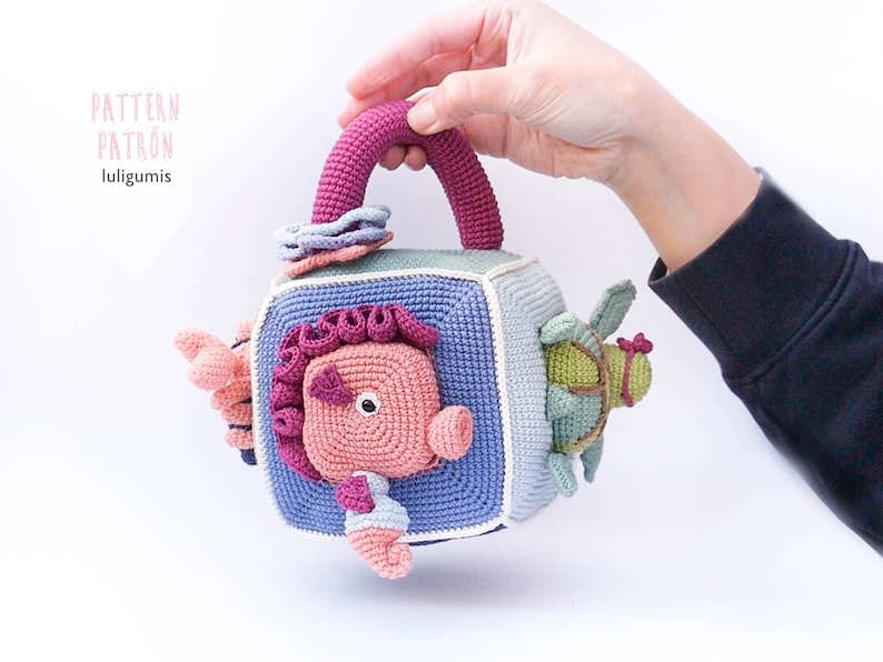 May include: A crocheted cube toy with a handle, featuring a pink seahorse, a green turtle, and a blue and white flower. The cube is made of different colored yarn and has a textured surface.