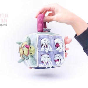 May include: A crocheted cube toy with a handle, featuring a turtle, jellyfish, and crab. The cube is made in shades of blue, green, purple, and pink.