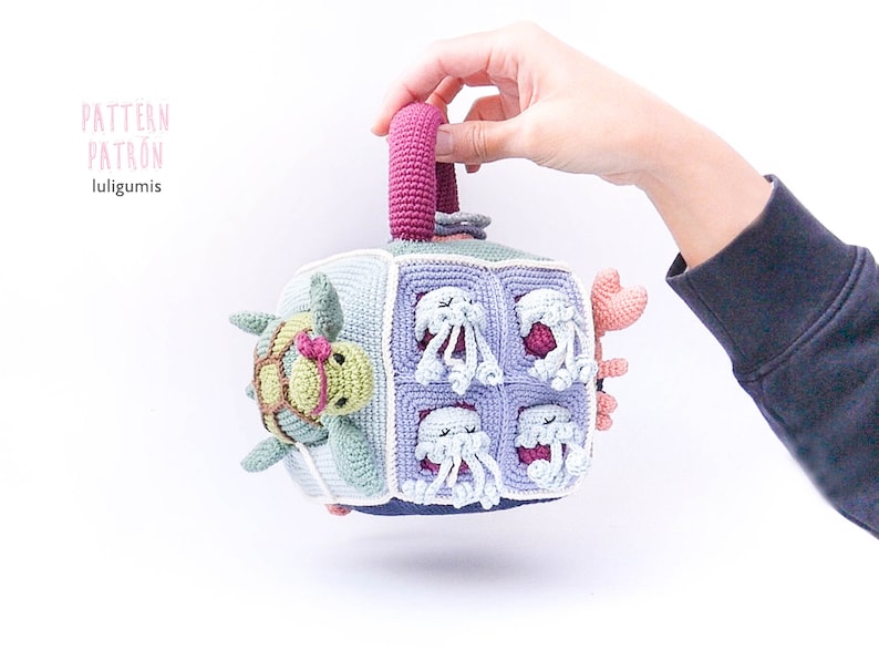 May include: A crocheted cube toy with a handle, featuring a turtle, jellyfish, and crab. The cube is made in shades of blue, green, purple, and pink.