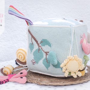 May include: A handmade crochet cube toy in light blue, featuring a tree branch design with green leaves. The cube has various attached crochet elements, including a snail, flower, and a rainbow handle. The text "PATTERN PATRON luligumis" is visible.