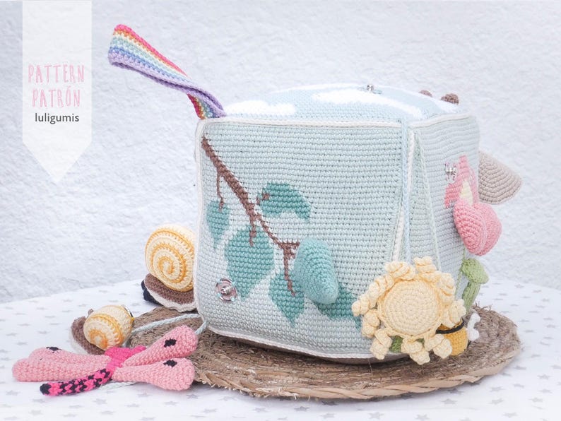 May include: A handmade crochet cube toy in light blue, featuring a tree branch design with green leaves. The cube has various attached crochet elements, including a snail, flower, and a rainbow handle. The text "PATTERN PATRON luligumis" is visible.