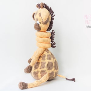 May include: A crocheted giraffe toy with brown spots and a brown mane. The giraffe is wearing a yellow crocheted scarf. The toy is sitting on a white background. The text "PATTERN PATRÓN luluigumis" is in the upper right corner of the image.