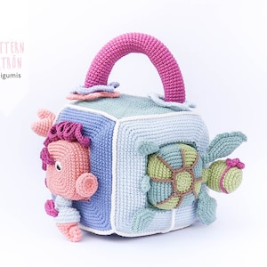 May include: A crocheted cube toy with a pink handle, featuring a turtle, a fish, and a flower. The cube is made of blue, green, and white yarn.