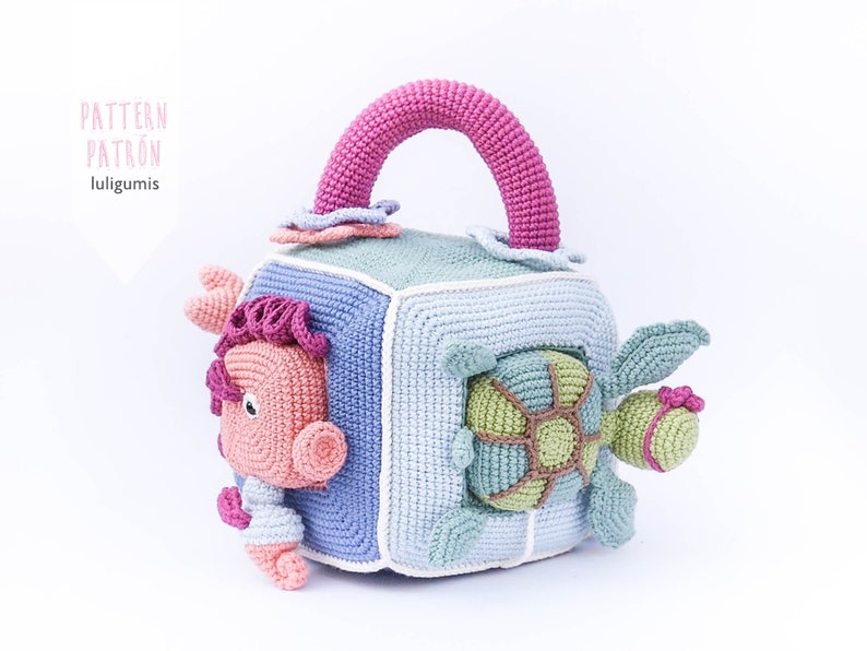 May include: A crocheted cube toy with a pink handle, featuring a turtle, a fish, and a flower. The cube is made of blue, green, and white yarn.