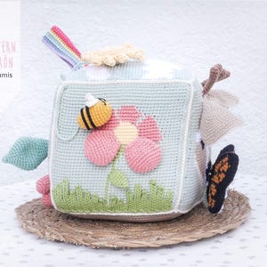May include: Crocheted baby toy cube with a flower, bee, and butterfly design. The cube is light blue with a flower in pink and yellow. A rainbow and other shapes are attached to the cube. The text "PATTERN PATRON luligumis" is on the side.
