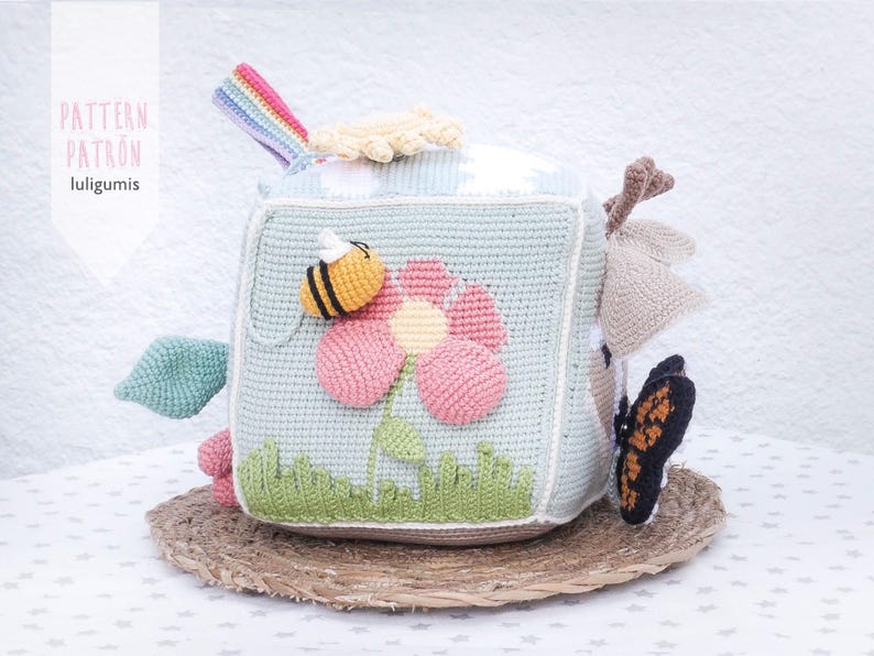 May include: Crocheted baby toy cube with a flower, bee, and butterfly design. The cube is light blue with a flower in pink and yellow. A rainbow and other shapes are attached to the cube. The text "PATTERN PATRON luligumis" is on the side.