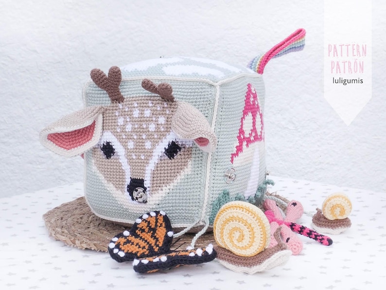 May include: A handmade crochet cube toy featuring a deer face, a mushroom, and a rainbow handle. The cube is light blue and beige, with brown antlers and pink accents. Includes a crochet butterfly and snails.