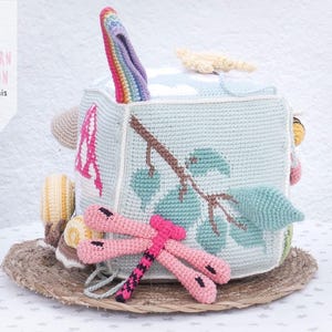 May include: A handmade crochet cube toy with various textures and colors. The cube features a rainbow-colored handle, a branch and leaf design, and a pink dragonfly. The text "PATTERN PATR&Oacute;N luligumis" is visible.
