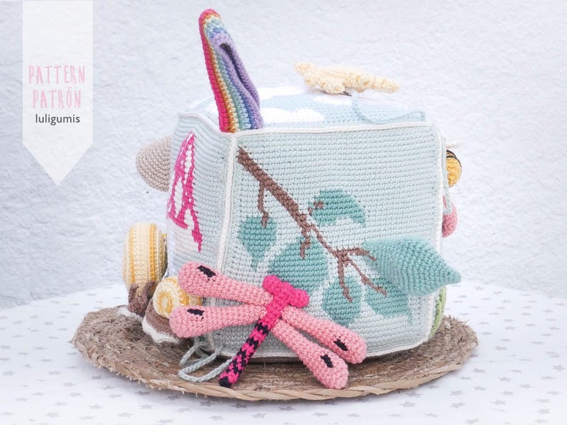 May include: A handmade crochet cube toy with various textures and colors. The cube features a rainbow-colored handle, a branch and leaf design, and a pink dragonfly. The text "PATTERN PATR&Oacute;N luligumis" is visible.