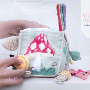 May include: A crocheted baby activity cube with a mushroom design in pink and white on a light blue background. The cube has various attached toys, including a snail, a butterfly, and a rainbow handle. The text "PATTERN PATRON luligumis" is visible.