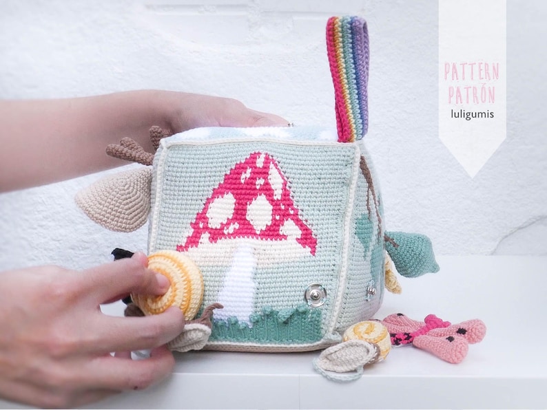 May include: A crocheted baby activity cube with a mushroom design in pink and white on a light blue background. The cube has various attached toys, including a snail, a butterfly, and a rainbow handle. The text "PATTERN PATRON luligumis" is visible.